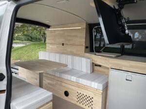 Petit van aménagé par Karr Koncept : banquette et table en hévéa massif face au réfrigérateur, porte latérale ouverte