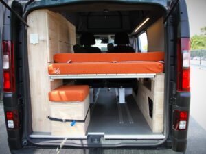 Van 4 places 4 couchages par Karr Koncept : le couchage haut orange et un coffre en hévéa massif, vus par les portes arrière