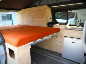 Van 4 places 4 couchages par Karr Koncept : le lit déplié aux matelas orange à côté du meuble cuisine en hévéa massif