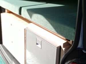 Van 7 places aménagé par Karr Koncept : façade grise du réfrigérateur logé sous le couchage, caisson en hévéa massif